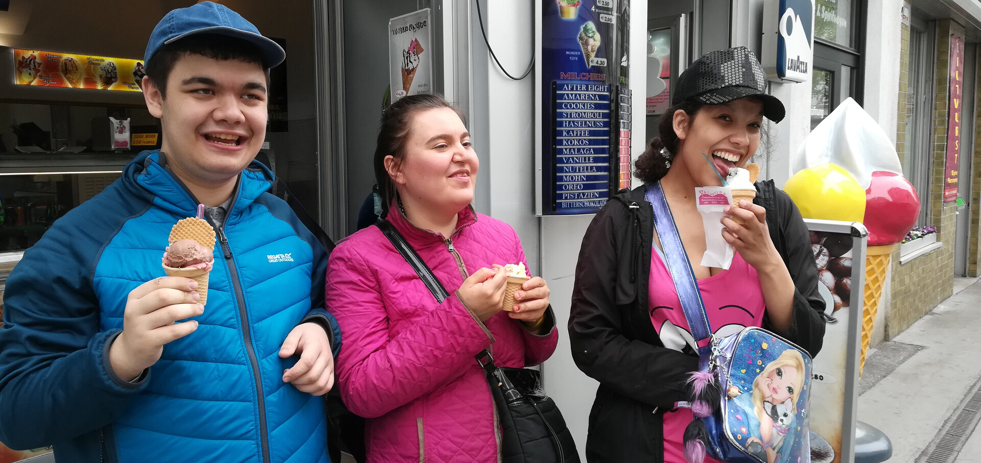 Drei junge Menschen stehen gut gelaunt vor einem Eissalon und genie&szlig;en ihr Eis in Waffeln. Links ein junger Mann in einer blauen Steppjacke und blauer Kappe, in der Mitte eine Frau in einer pinken Jacke und rechts eine junge Frau mit schwarzer Kappe und bunter Umh&auml;ngetasche, die gerade an ihrem Softeis schleckt. Im Hintergrund sind die Eiskarte und eine gro&szlig;e Werbefigur in Form einer Eist&uuml;te zu sehen.