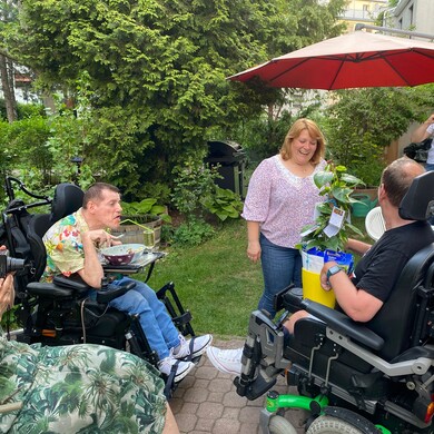 Mehrere Menschen mit und ohne Behinderung sitzen und stehen in einem Garten zusammen. Eine Frau bekommt eine Topfpflanze als Geschenk &uuml;berreicht.