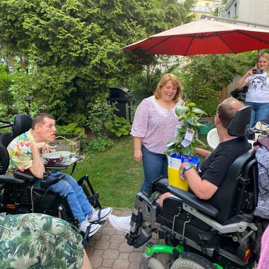 Eine fr&ouml;hliche Gruppe von Personen im Garten der Wohngemeinschaft B&ouml;ckh an einem sonnigen Tag. Im Zentrum &uuml;berreicht eine l&auml;chelnde Frau einem Mann, der in einem elektrischen Rollstuhl sitzt, eine gro&szlig;e Gr&uuml;npflanze in einer gelben Tasche. Ein weiterer Mann im Rollstuhl beobachtet die Szene beim Essen an einem kleinen Tisch-Aufsatz. Im Hintergrund steht eine junge Frau unter einem roten Sonnenschirm und macht ein Foto. Der Garten ist dicht begr&uuml;nt und wirkt einladend und gemeinschaftlich