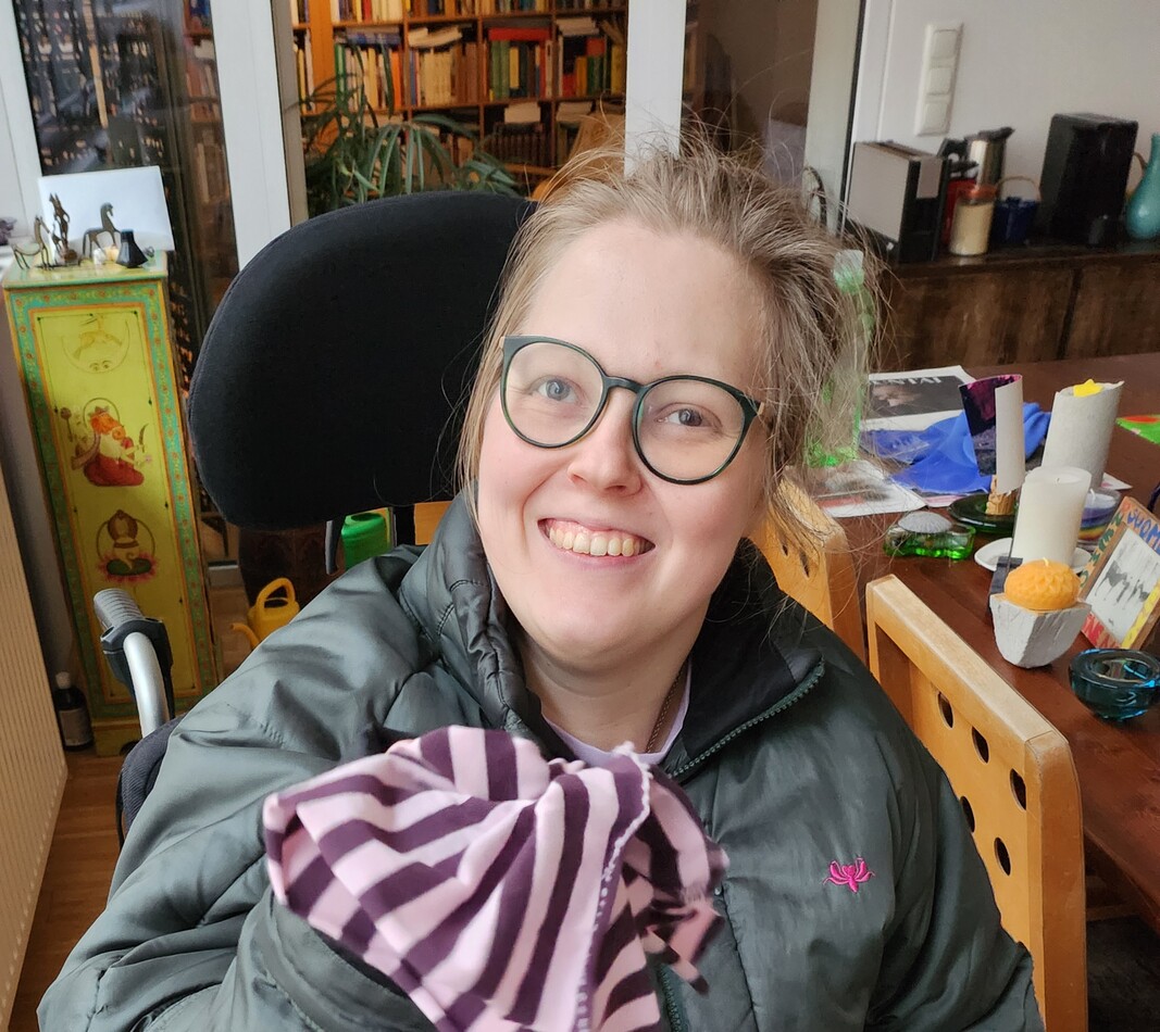 Aktuelles-Spendenaufruf-Hebelifter Liisa lächelt fröhlich in die Kamera. Sie trägt eine Brille, eine dunkle Jacke und schwarze Hose. In der rechten Hand hält sie ein gestreiftes Geschirrtuch. Sie sitzt in einem schwarz-orangen Rollstuhl.
