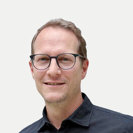 Portr&auml;taufnahme von Mag. David Brauner, Leitung der Tagesstruktur SoHo bei BALANCE. Er blickt freundlich l&auml;chelnd in die Kamera, tr&auml;gt eine markante schwarze Brille und ein dunkles Hemd. Das Bild zeigt ihn vor einem neutralen, hellen Hintergrund.