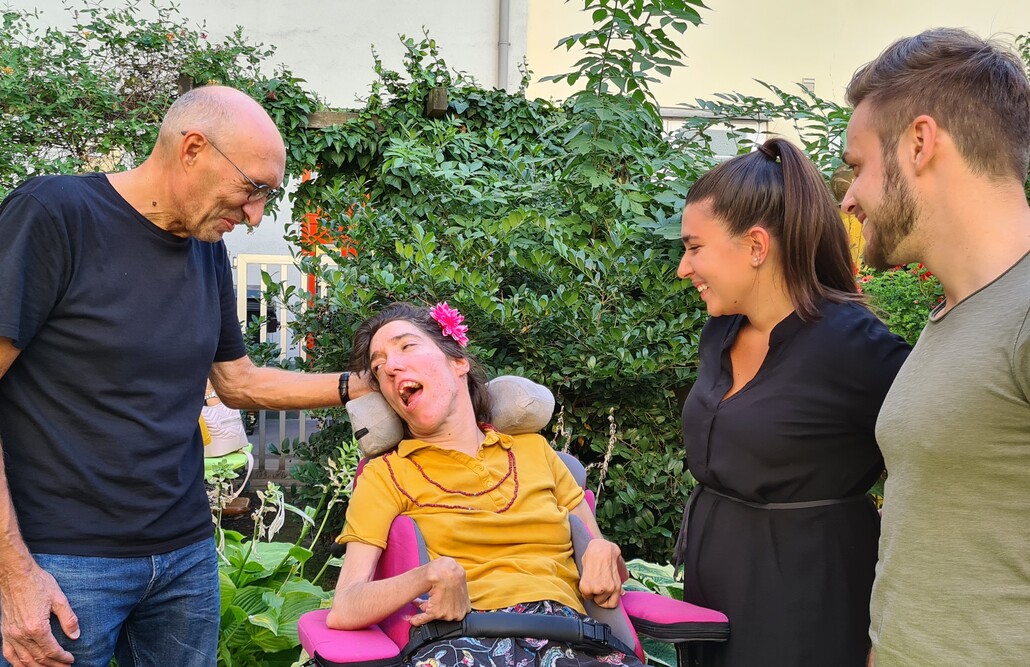 Eine Frau im Rollstuhl sitzt in einem gr&uuml;nen Garten und wird von drei Personen umgeben, die sich ihr zugewandt unterhalten und l&auml;cheln.