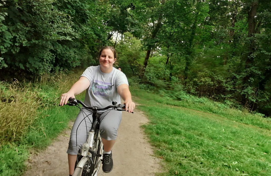 Eine junge Frau f&auml;hrt l&auml;chelnd mit dem Fahrrad auf einem Weg neben einer gr&uuml;ne Wiese. Im Hintergrund sieht man B&auml;ume.