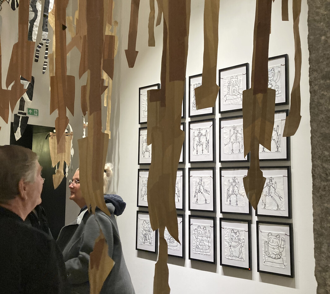 Durch herabh&auml;ngende Teile von Karton-Figuren sieht man 20 Zeichnungen in einem Rechteck angeordnet. Zwei Besucher stehen davor und betrachten diese Collage.