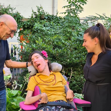 Eine Frau im Rollstuhl sitzt in einem grünen Garten und wird von drei Personen umgeben, die sich ihr zugewandt unterhalten und lächeln.