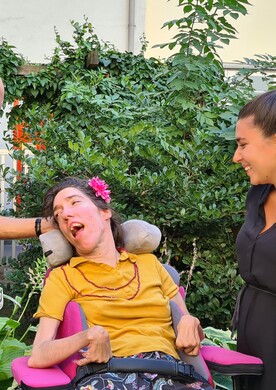 Eine Frau im Rollstuhl sitzt in einem gr&uuml;nen Garten und wird von drei Personen umgeben, die sich ihr zugewandt unterhalten und l&auml;cheln.