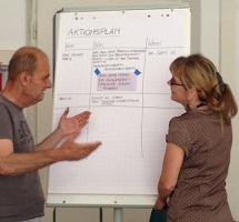 links ein Mann rechts eine Frau zwischen ihnen ein Flipchart