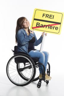 Frau im Rollstuhl sitzend hält ein gelbes Schild mit schwarzem Trennstrich in der Mitte. Oben steht frei, im unteren Teil Barrieren