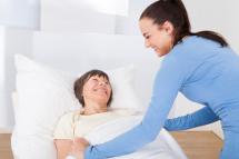 im Bett liegende lächelnde Frau wird von einer anderen stehenden lächelnden Frau zugedeckt.